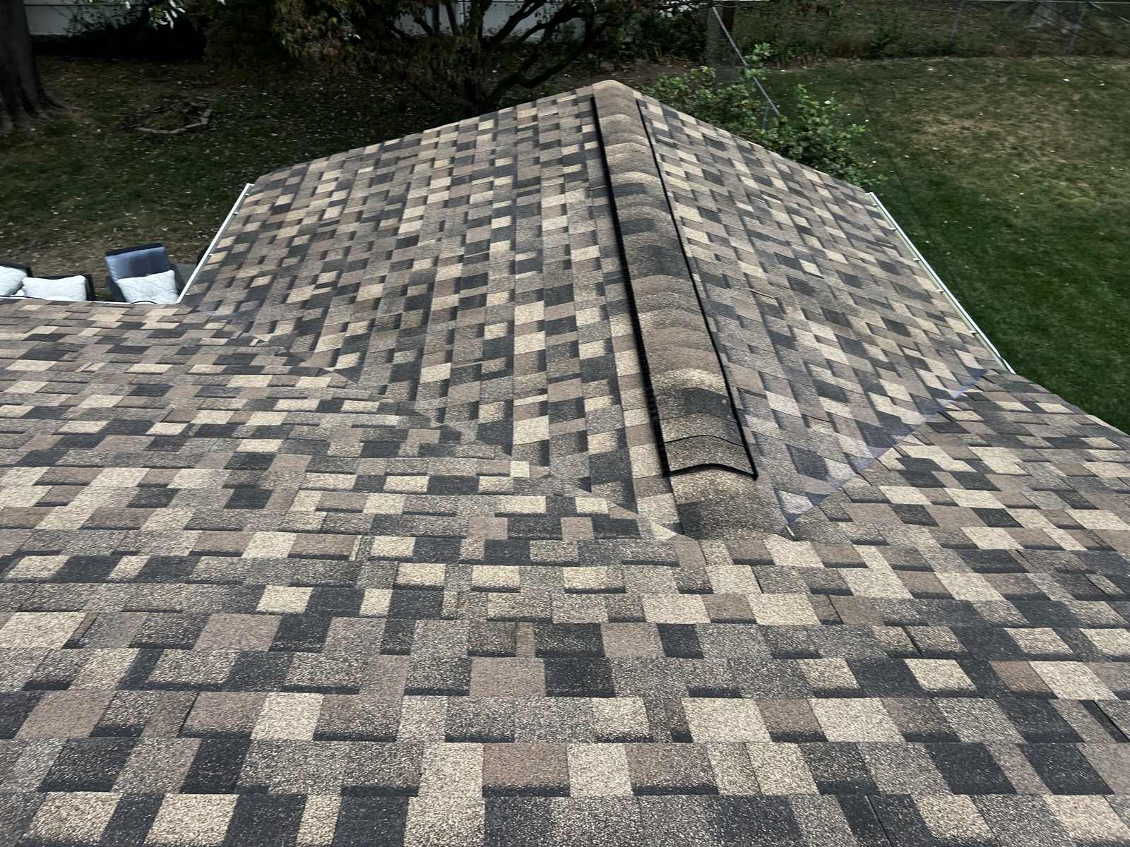 A finished hip roof photographed from above, laid in blended tan, grey and brown architectural shingles, with a black ridge vent running the peak and lawn and shrubbery visible past the eave