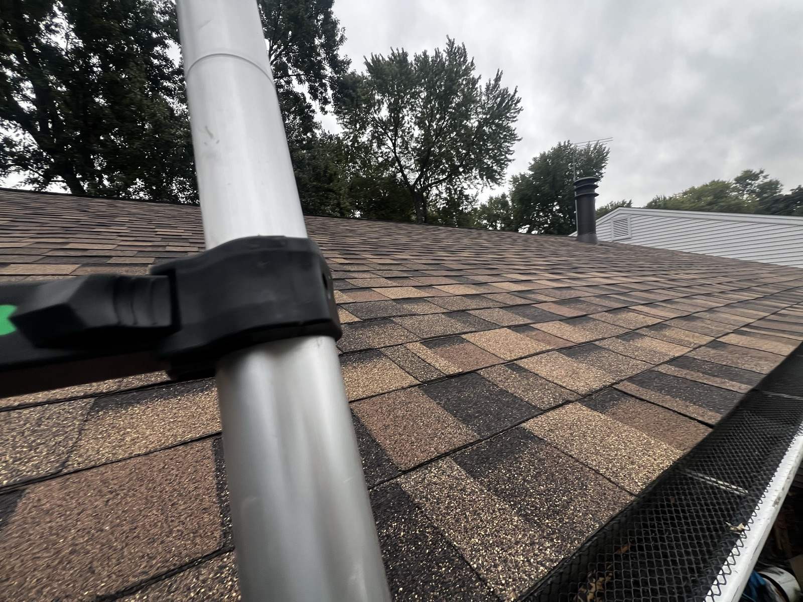 A ladder rail fitted with a black standoff bracket leaning across a field of new brown and tan shingles close to the eave, mesh gutter guard visible along the trough at the lower right, trees and a grey overcast sky above