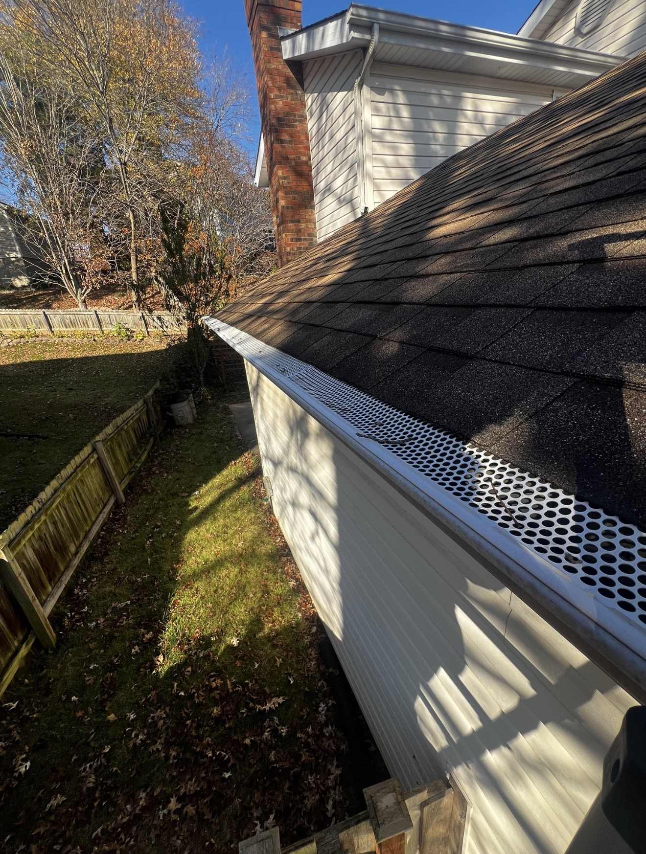 A run of white perforated aluminium gutter guard fitted along an eave of dark grey shingles, with a red brick chimney and white lap siding rising behind it and a leaf covered back garden inside a weathered wooden fence below