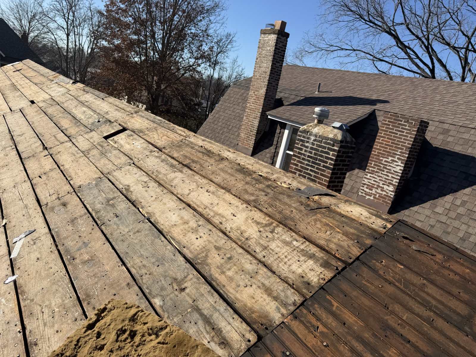 A roof stripped down to old board decking, the planks running diagonally with wide gaps between several of them, two brick chimneys and a metal turbine vent standing on the roof section beyond, bare winter branches and clear blue sky