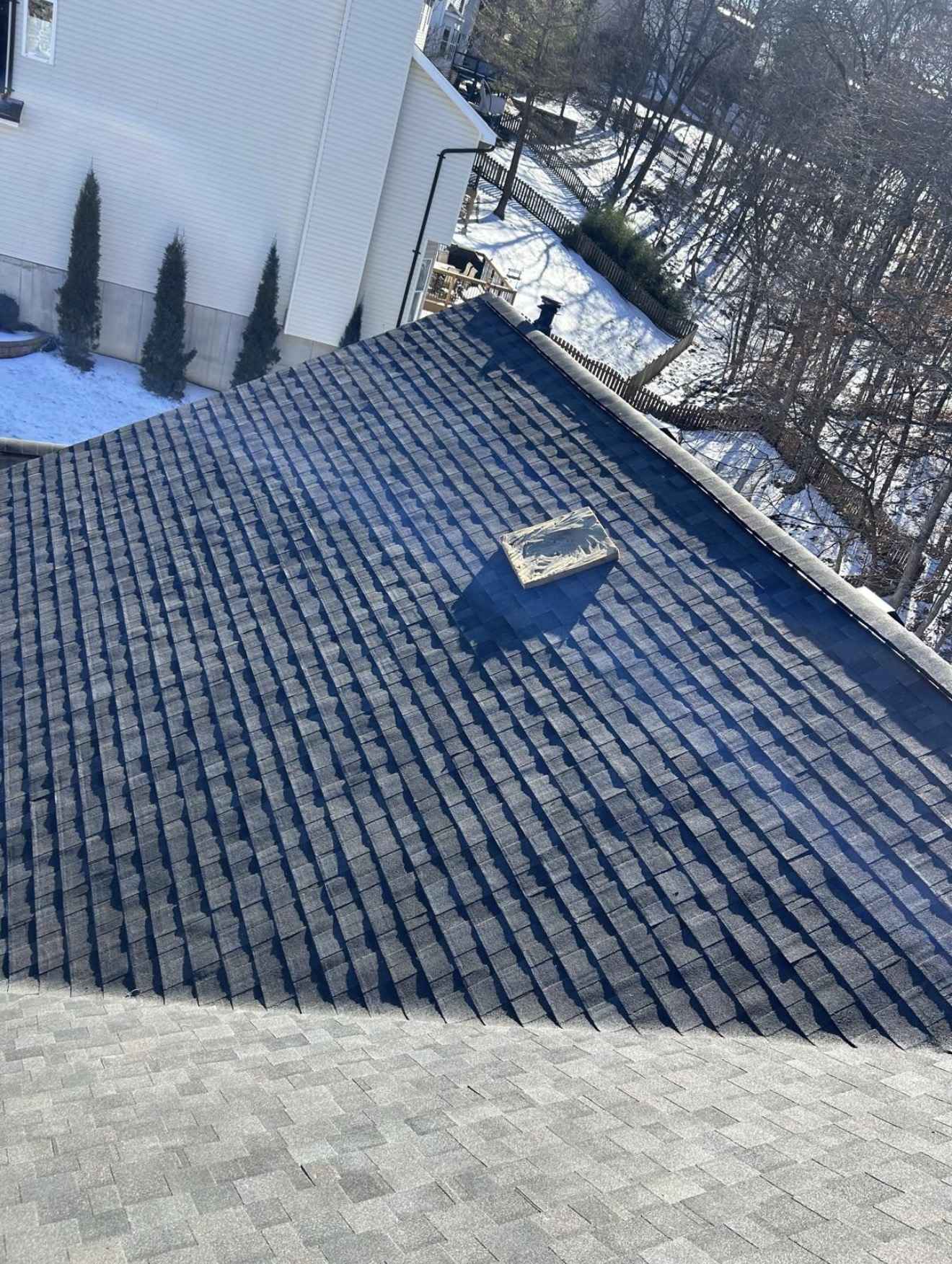 A large roof plane part way through reshingling, dark new courses running up the slope with a wrapper board left lying on it, a white lap sided wall and tall narrow evergreens to the left, snow covering the yard and a wooded ravine behind a dark metal fence