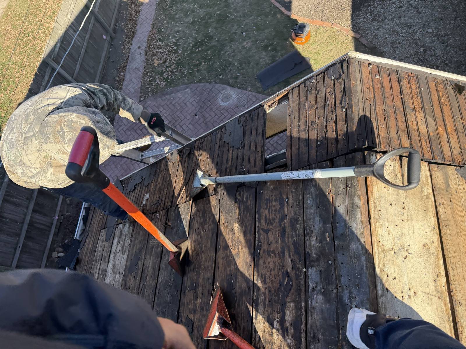 Two long handled tear off spades and a red flat bar lying on a roof stripped back to bare board decking, photographed from above, with a worker in a camouflage hooded jacket kneeling at the far edge and a brick paved patio on the ground below